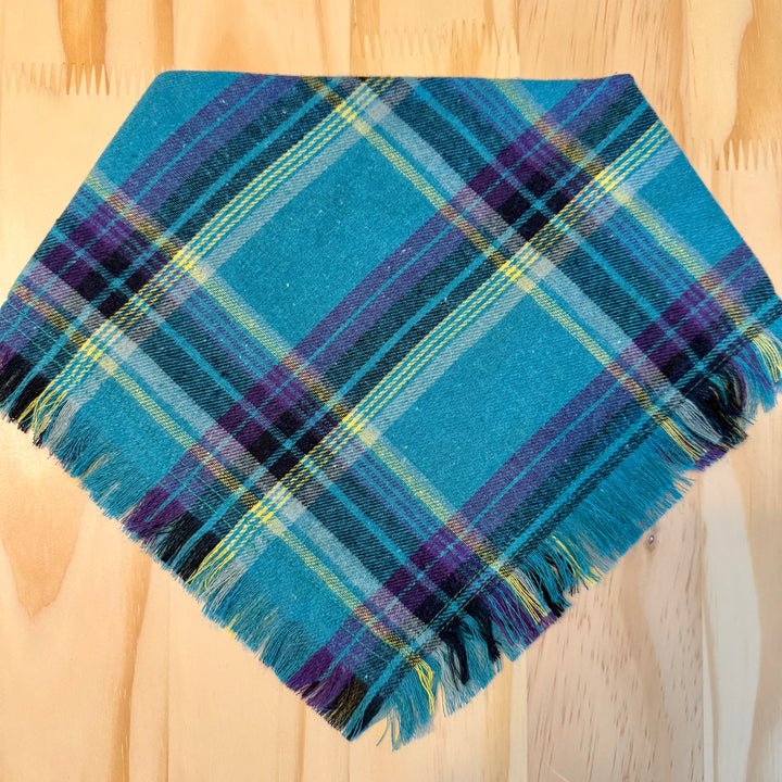 A multicolor plaid dog bandana laid flat on a light wood background.