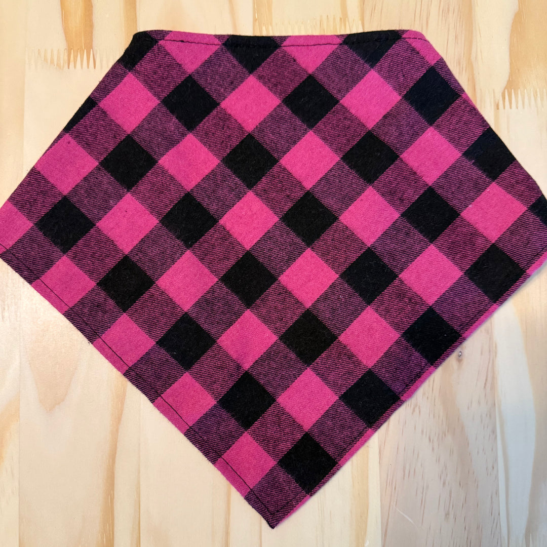 A pink and black plaid dog bandana laid flat on a light wood background.