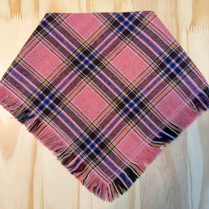 A pink multicolor plaid dog bandana laid flat on a light wood background.