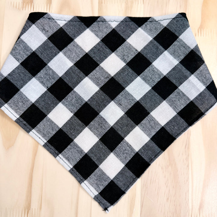 A black and white plaid dog bandana laid flat on a light wood background.